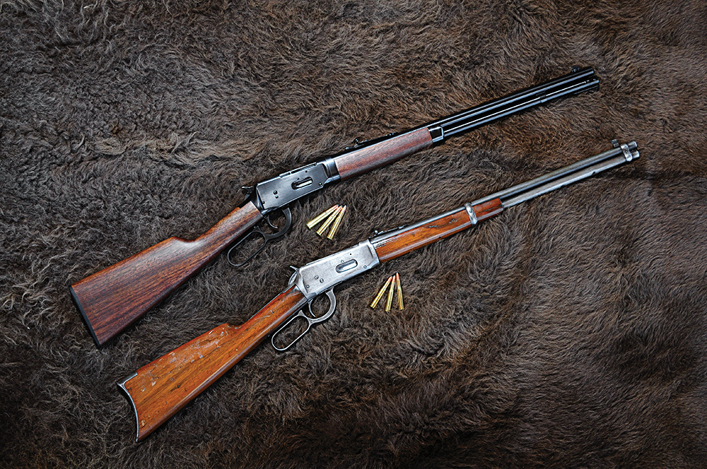 A pair of lever-action Winchester rifles, one old and worn, the other new, side by side on a dark brown hide.