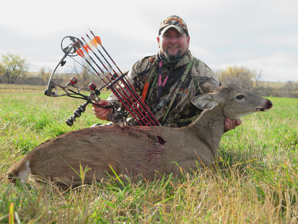 A bowhunter, holding a compound bow and arrow setup, seated behind a dead whitetail doe.