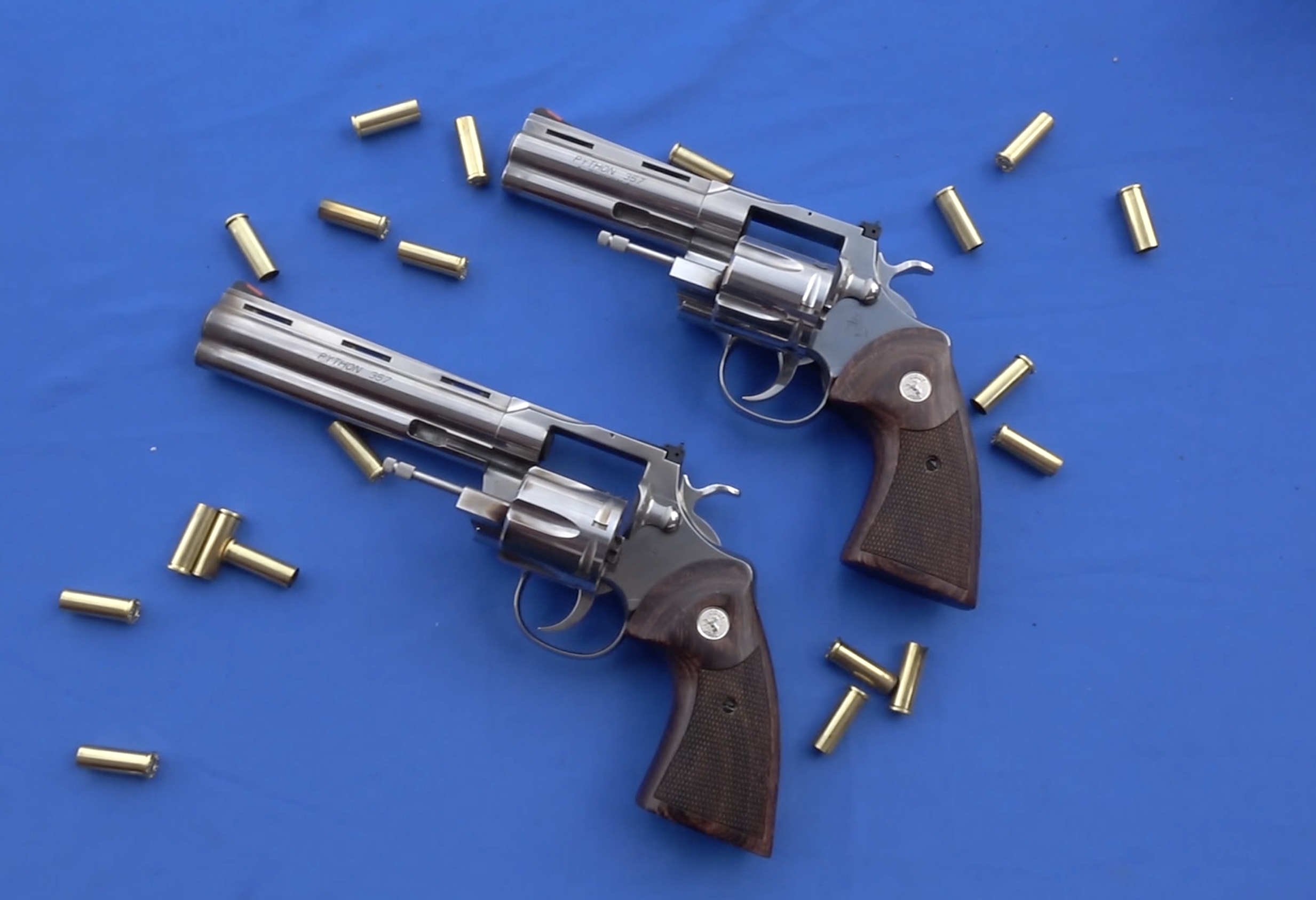 two Colt Python revolvers, one 6-inch barrel and one 4.25-inch barrel, with the wheels open on a blue background with empty cartridge cases