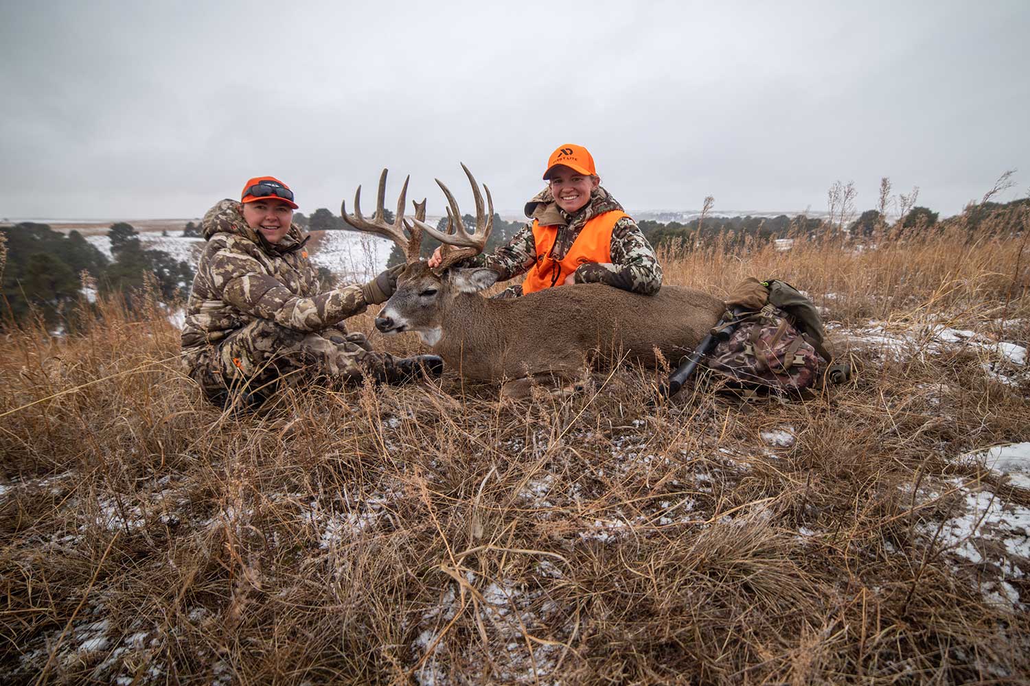Two hunters with a mule deer in the snow and grass.