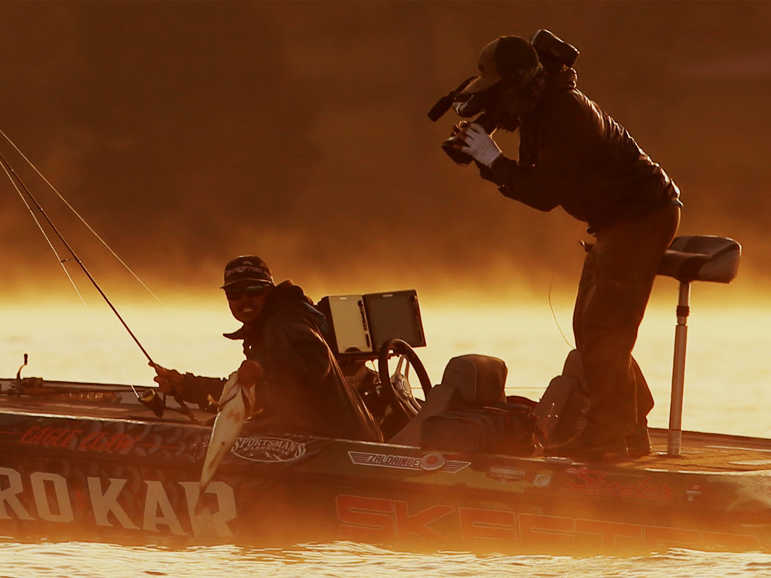 A bass angler pulling a fish into a boat while a camera man films.