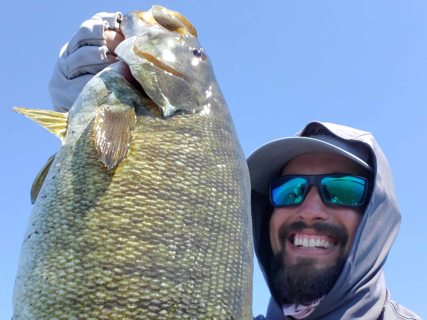 A angler in a cap and hoodie holds up a large smallmouth bass.