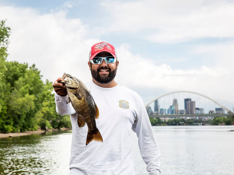 A man holding up a largemouth bass with a city in the background.