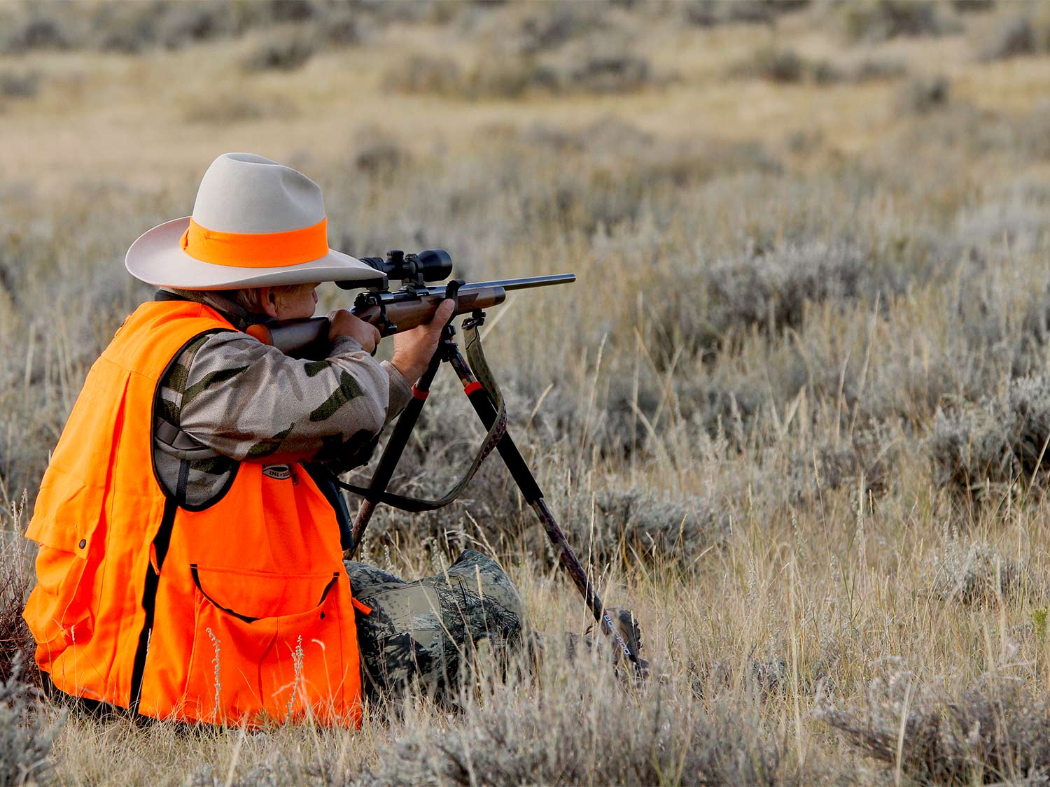A hunter in orange and camo aims a rifle in a field.