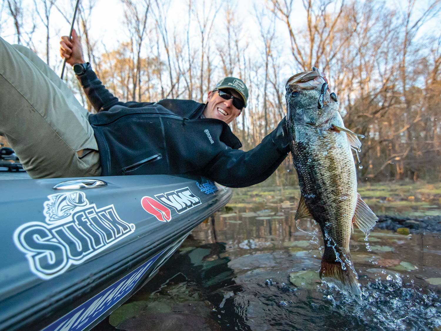 Patrick Walterns on the side of a boat hoisting a largemouth bass from the water.