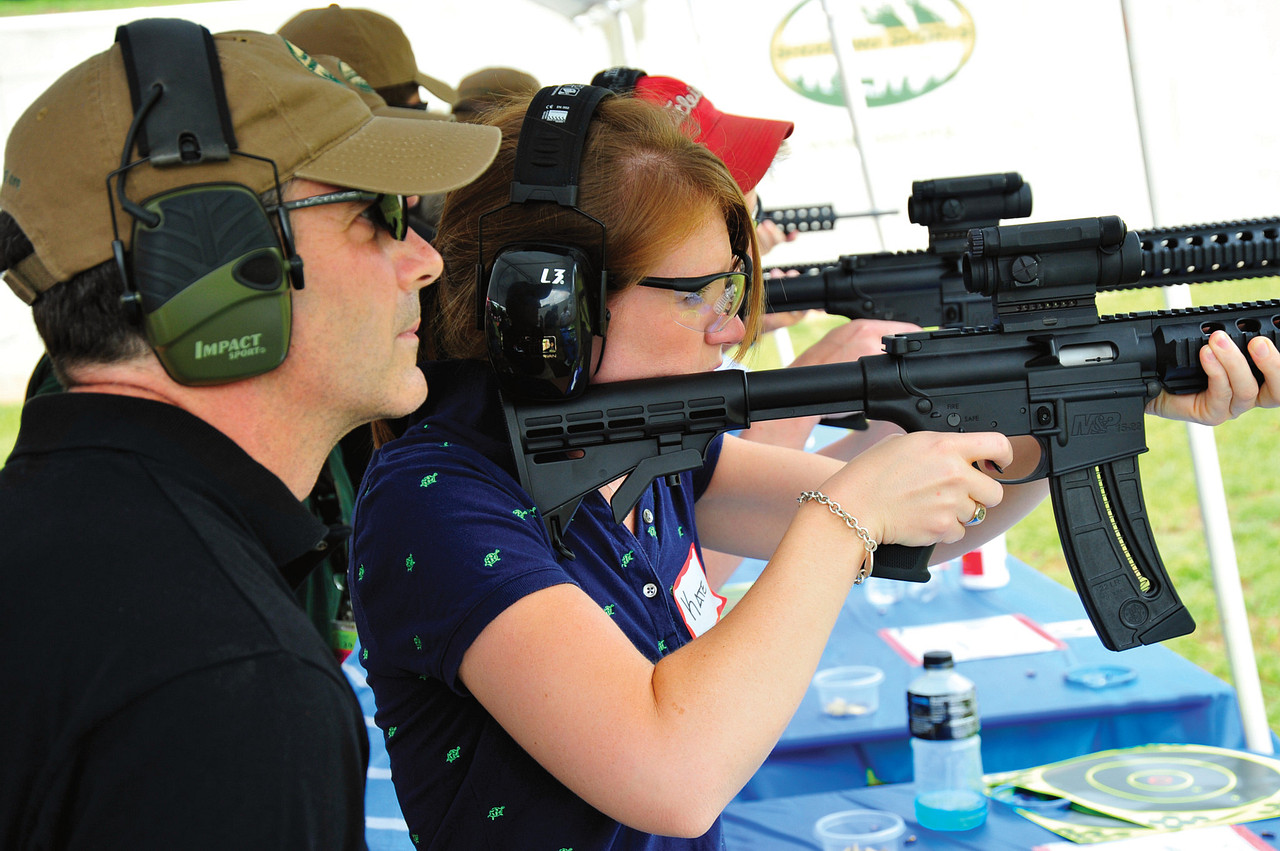 A woman holds a rifle to her should while taking aim, as an instructor stands beside her offering tips. Both are wearing shooting protection gear.