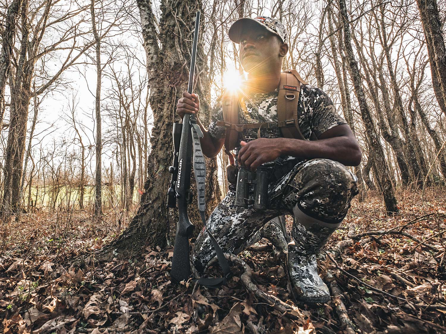 A young deer hunter kneels in the woods wearing full camo gear and holding a rifle in one hand.