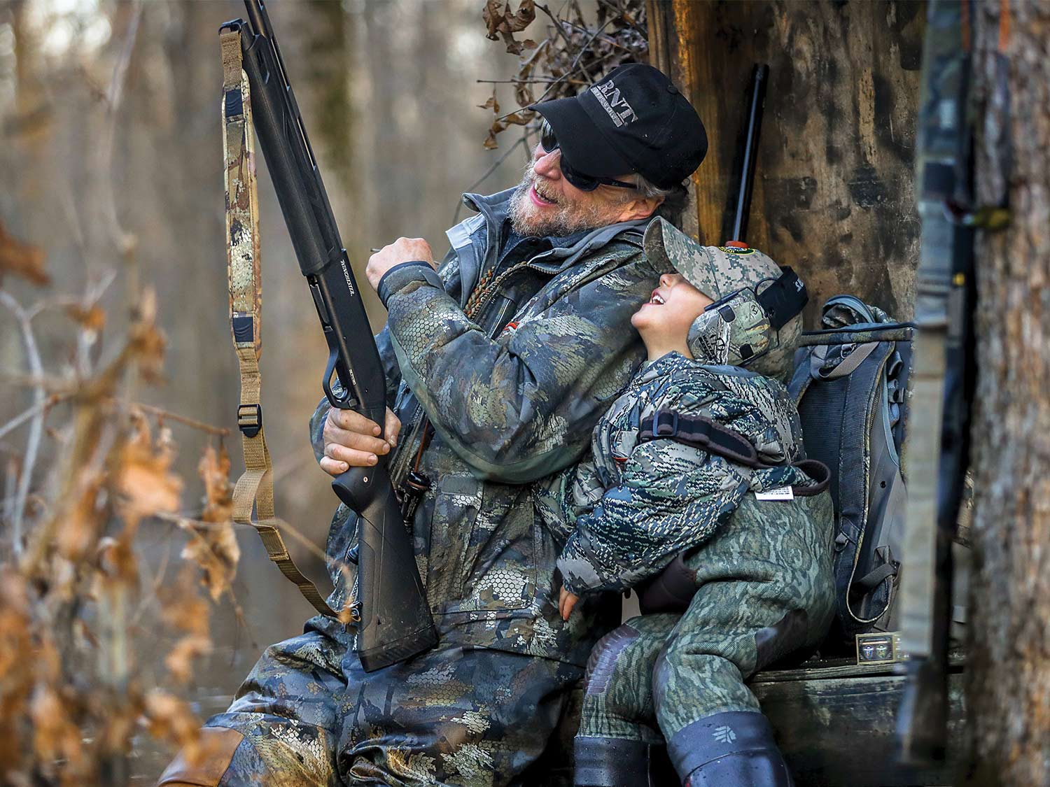 An elderly gentleman holding a rifle laughs and smiles with his grandchild in a tree stand.