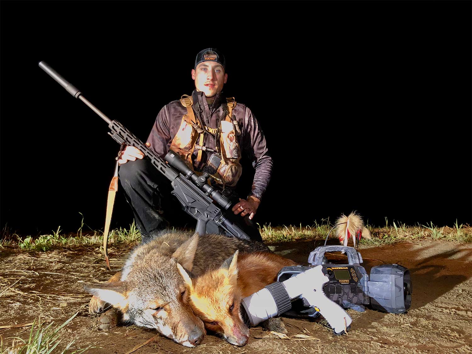 A hunter takes a knee behind a dead coyote and a fox at night with a rifle over his raised knee.