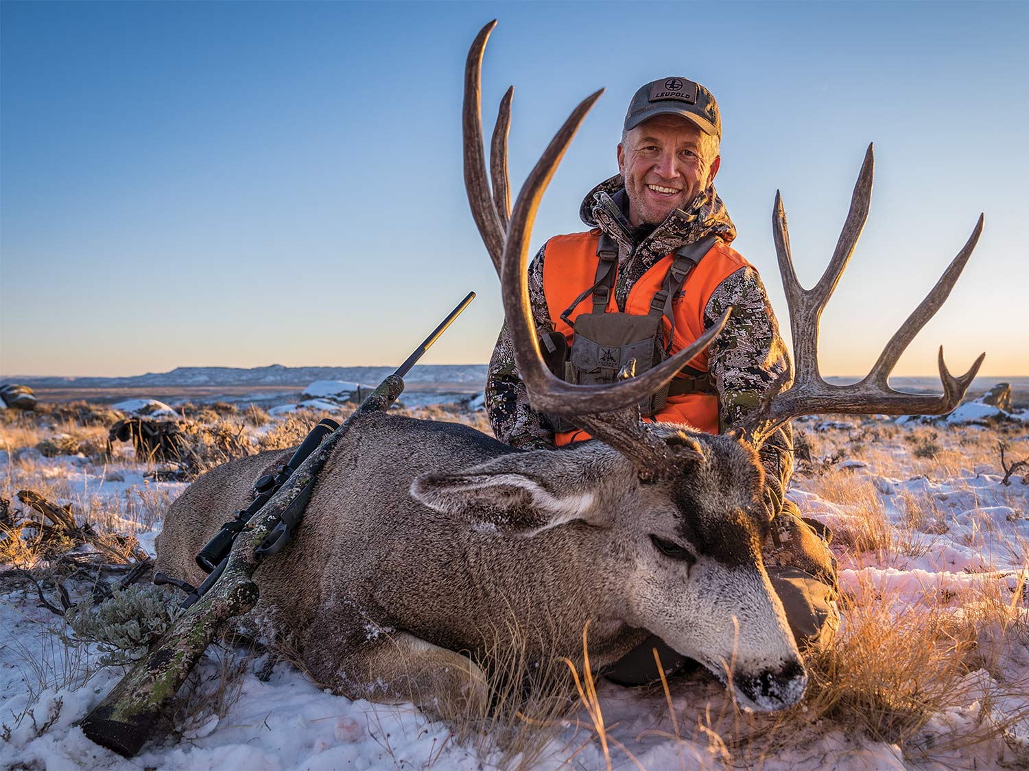 A hunter smiles and kneels behind a large mule deer in the snow.
