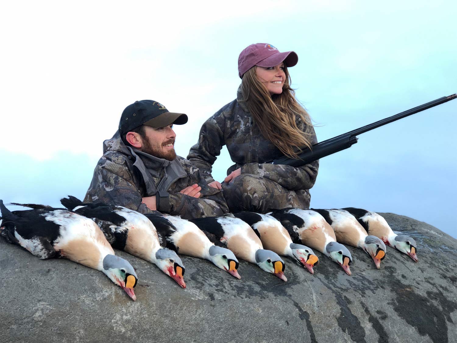 Two hunters, man and woman, sit on a rock next to a limit of king ducks.