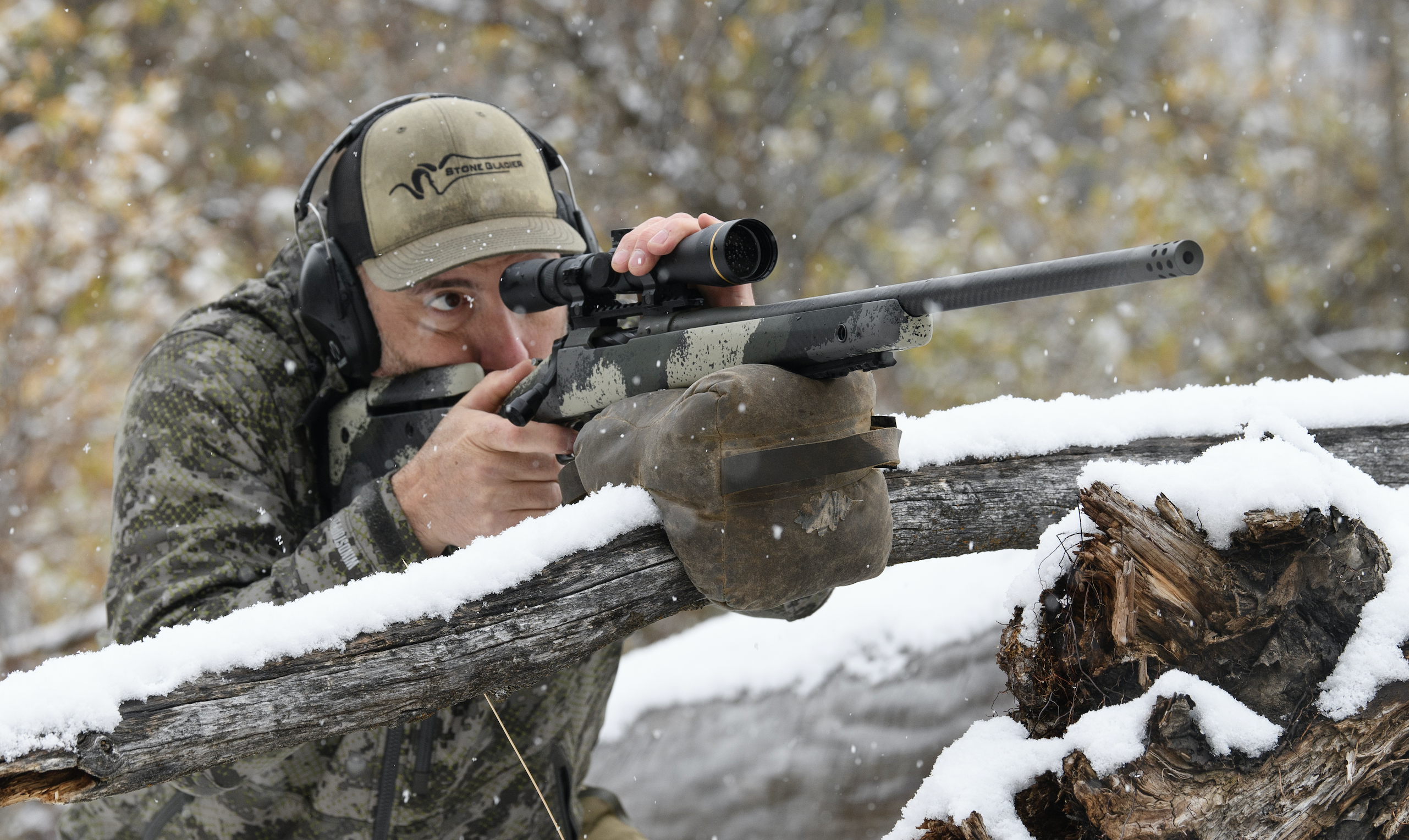 A hunter looks through the scope on his deer rifle, resting on a branch, in the snowy woods.