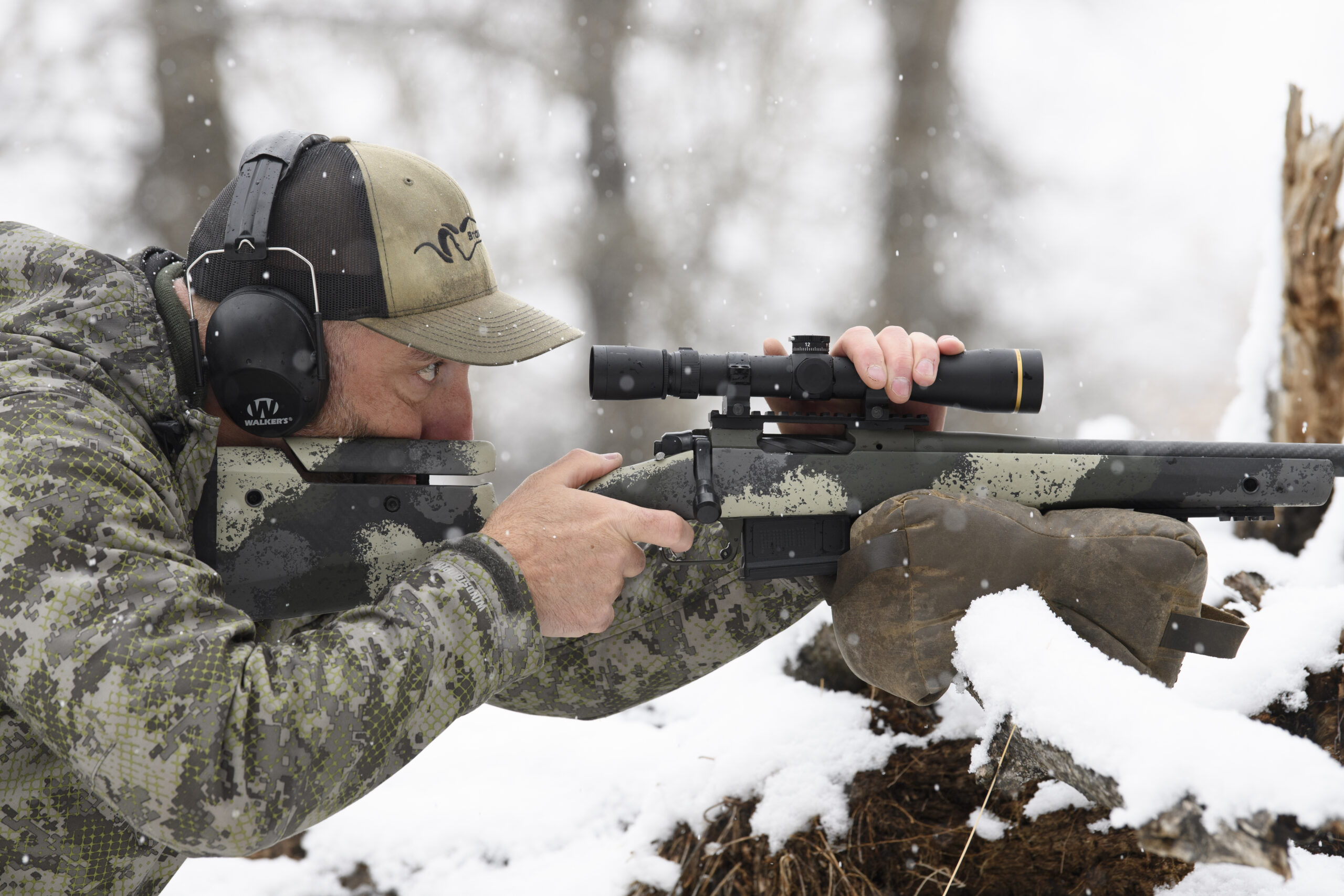 A hunter steadies his Springfield Waypoint bolt-action rifle on top of a shooting bag resting on a branch in a snowy field.