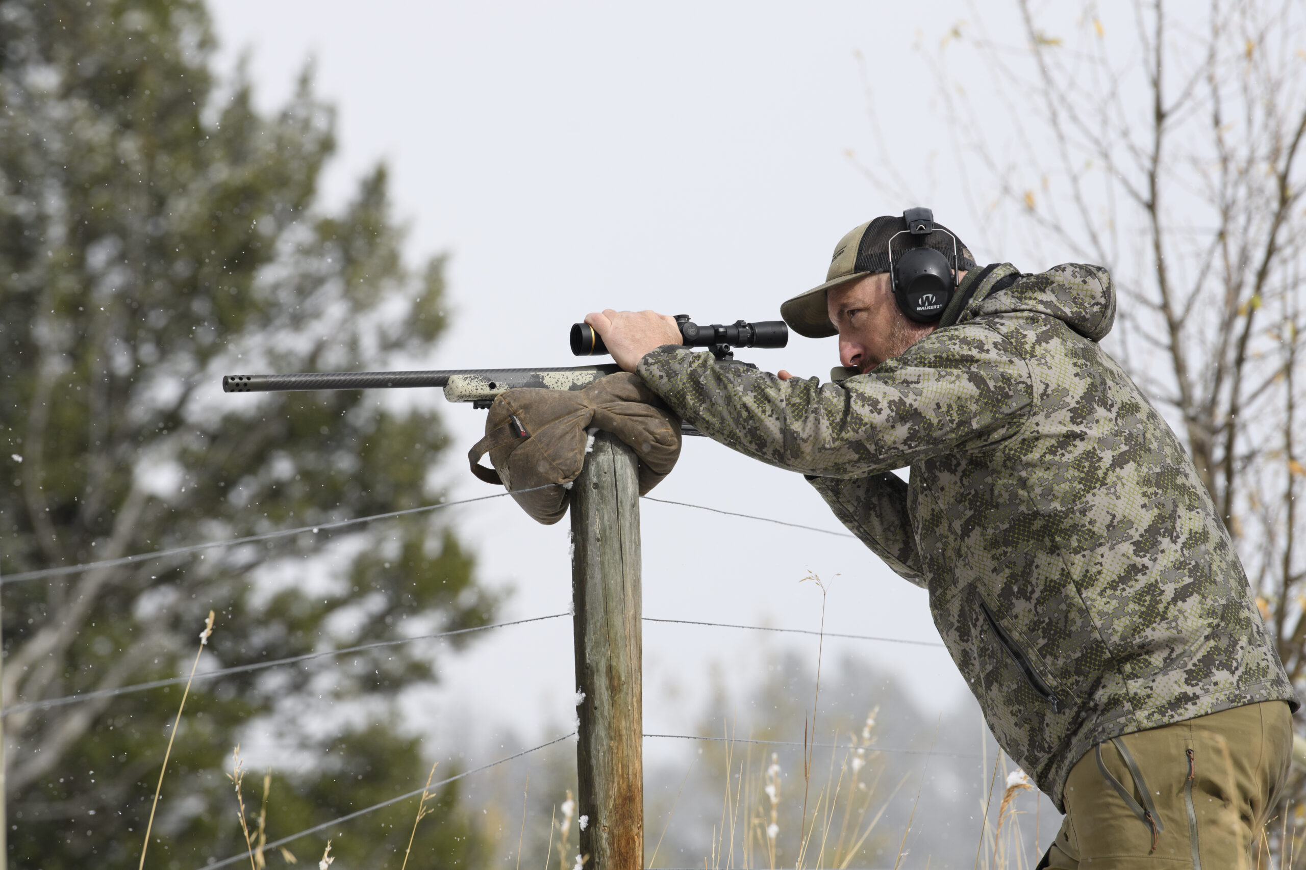 A Western hunter readies himself for a shot behind his bolt-action rifle, which is resting on a fence post.