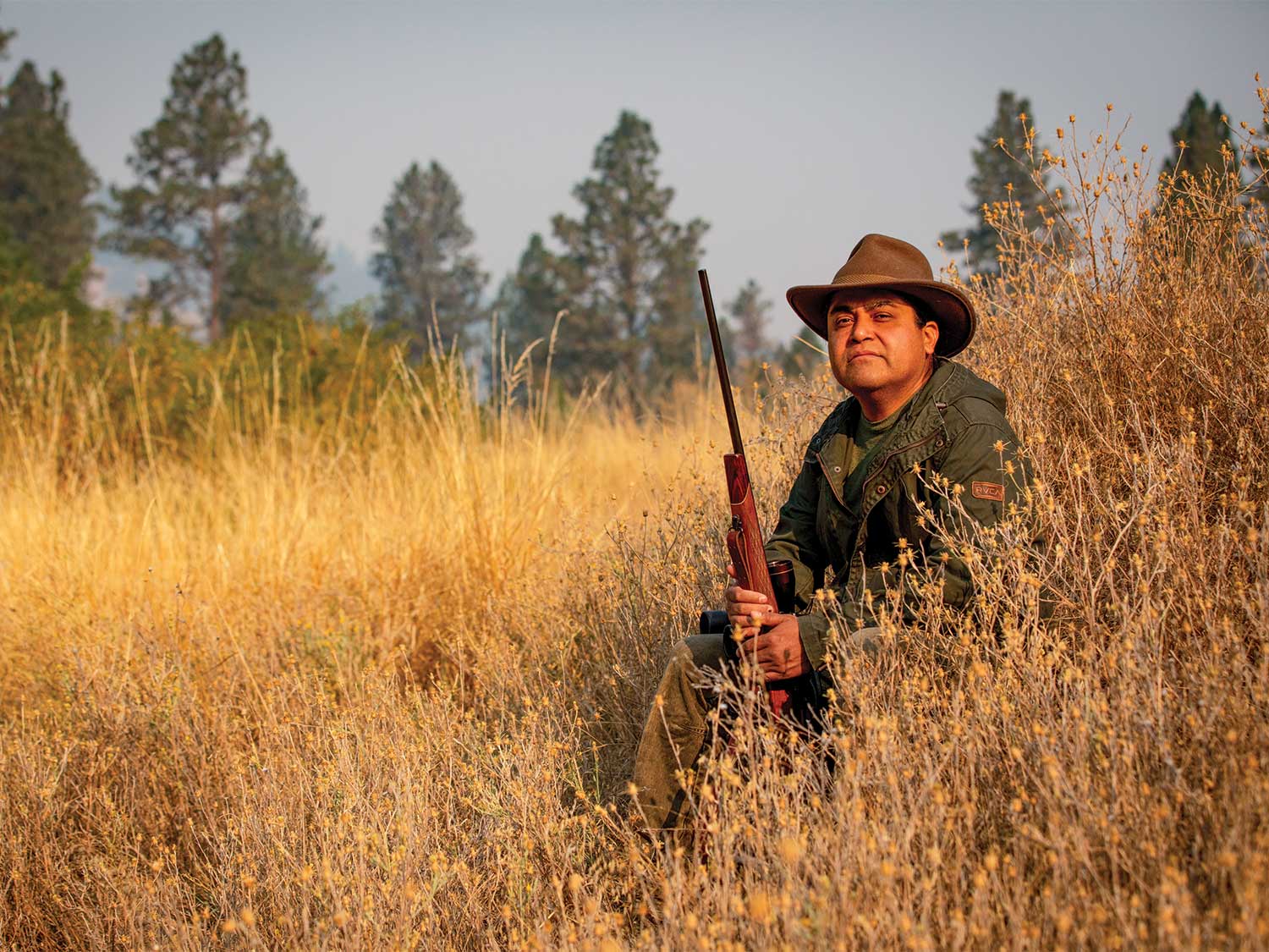 A native hunter, Josiah Pinkham, sits in a field of tall weeds and holds a rifle.