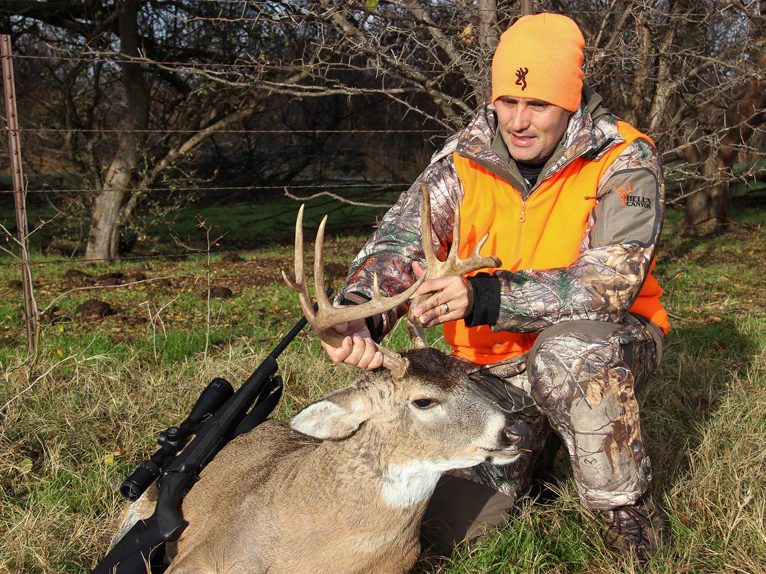 A hunter in camo and orange kneels next to a whitetail buck.