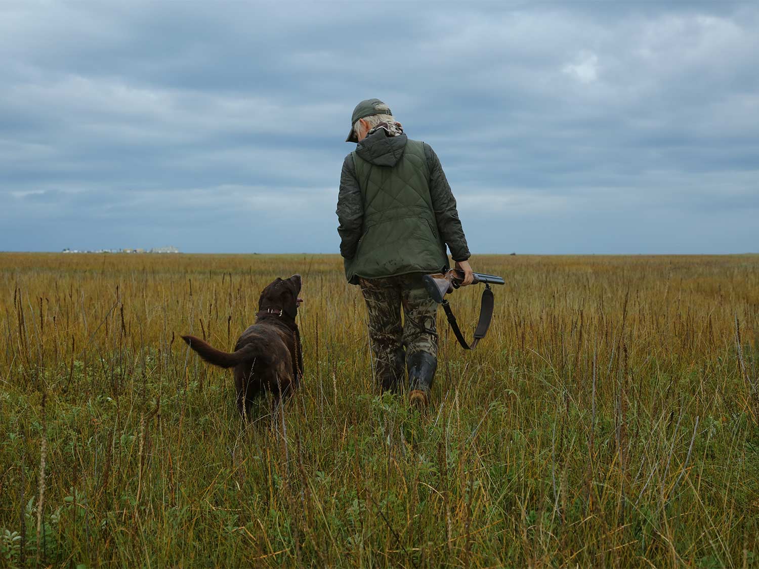 A hunter in camo and jacket holds a gun while walking through an open field. A hunting dog walks beside them.