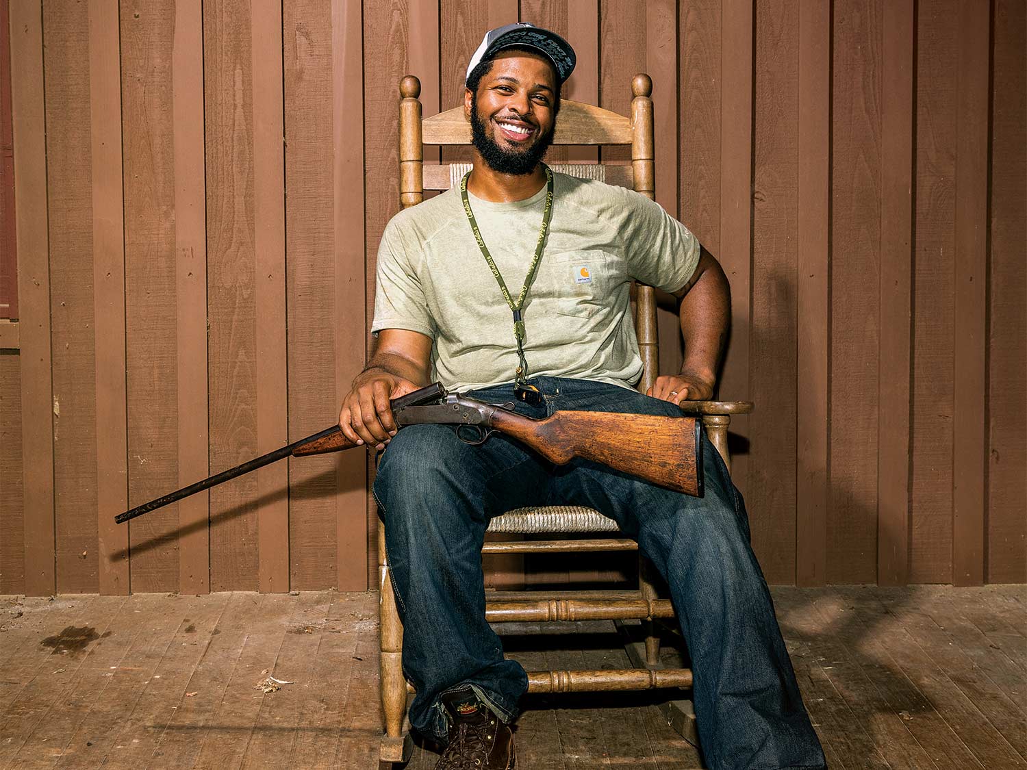 A hunter sits in a rocking chair, smiling, with a gun in his lap.