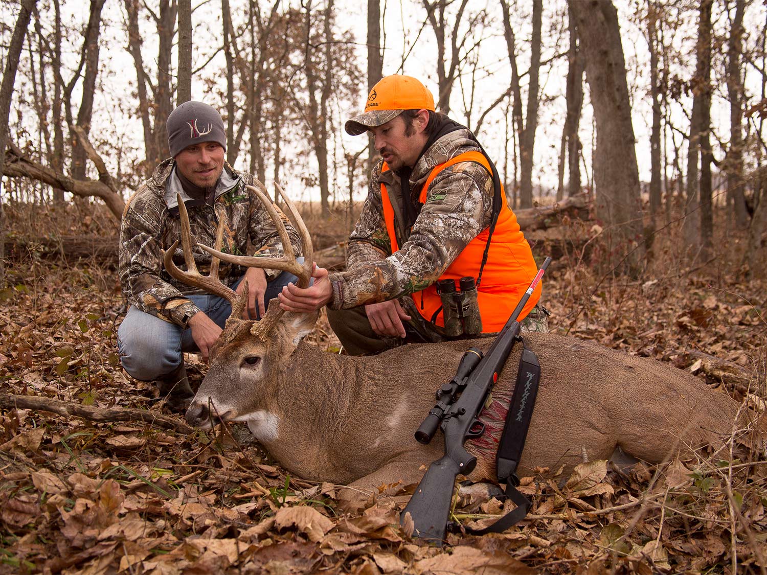 Two hunters kneel behind a deer and hold its head up by the antlers.