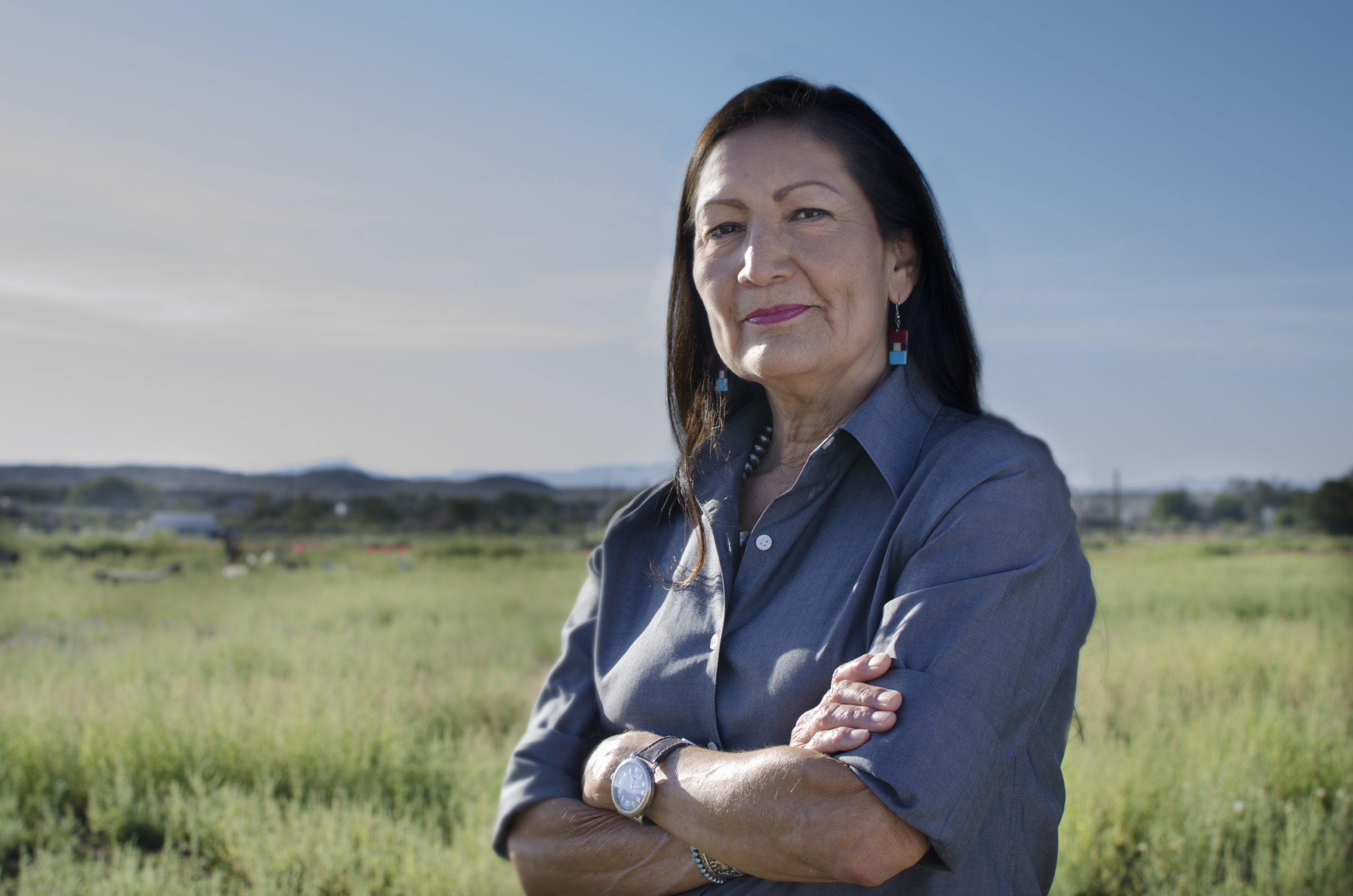 A New Mexican woman with dark hair and turquoise earrings crosses her arms and looks at the camera in an open field.