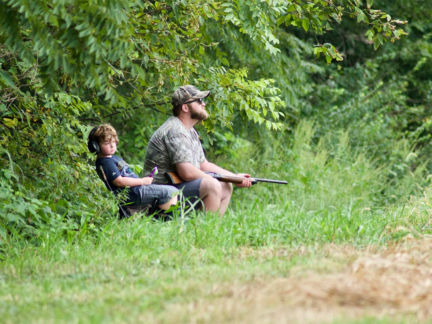 A man and a young kid sit at the edge of a field for hunting.