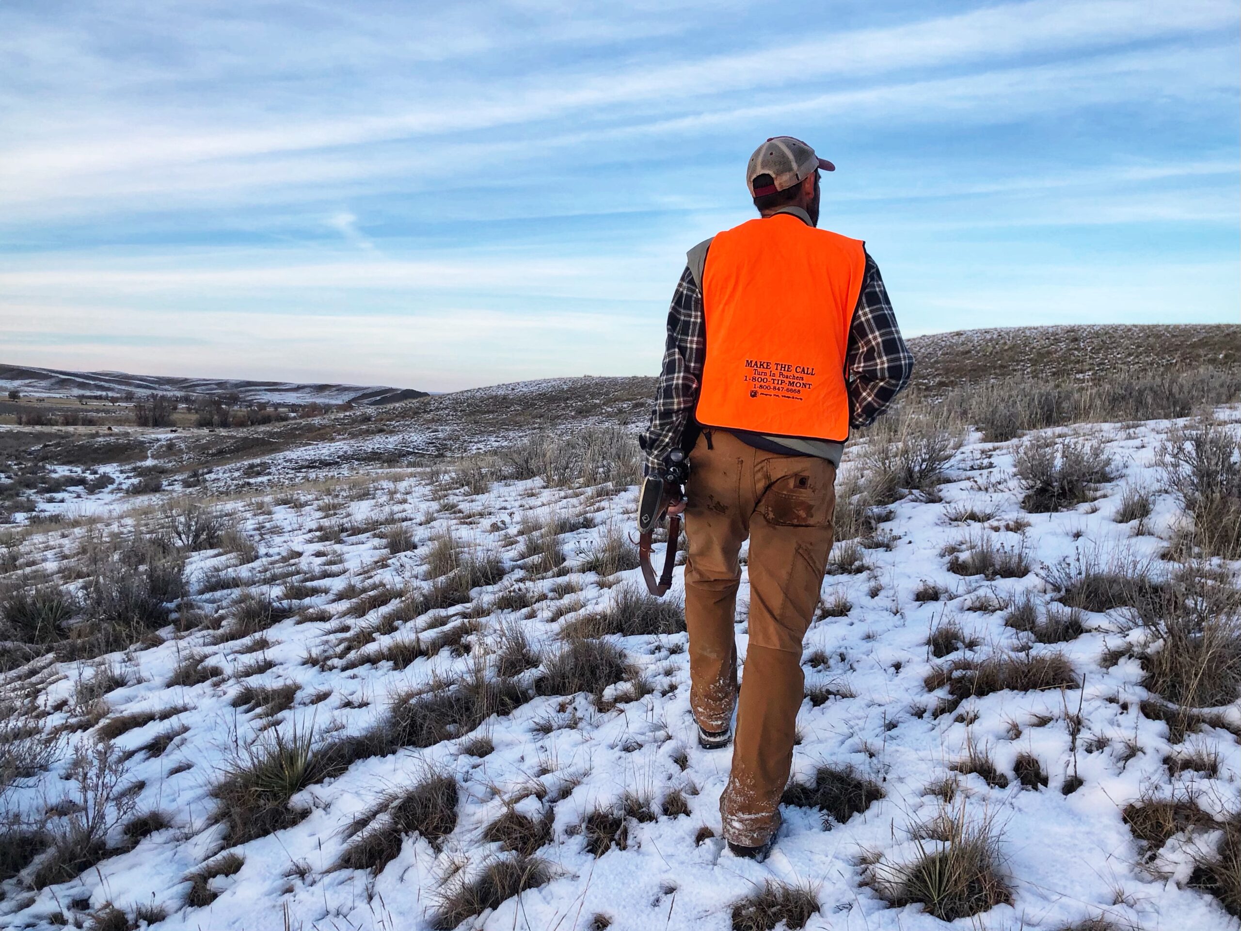A Western deer hunter in a flannel and blaze orange vest carries a rifle on the Montana prairie.