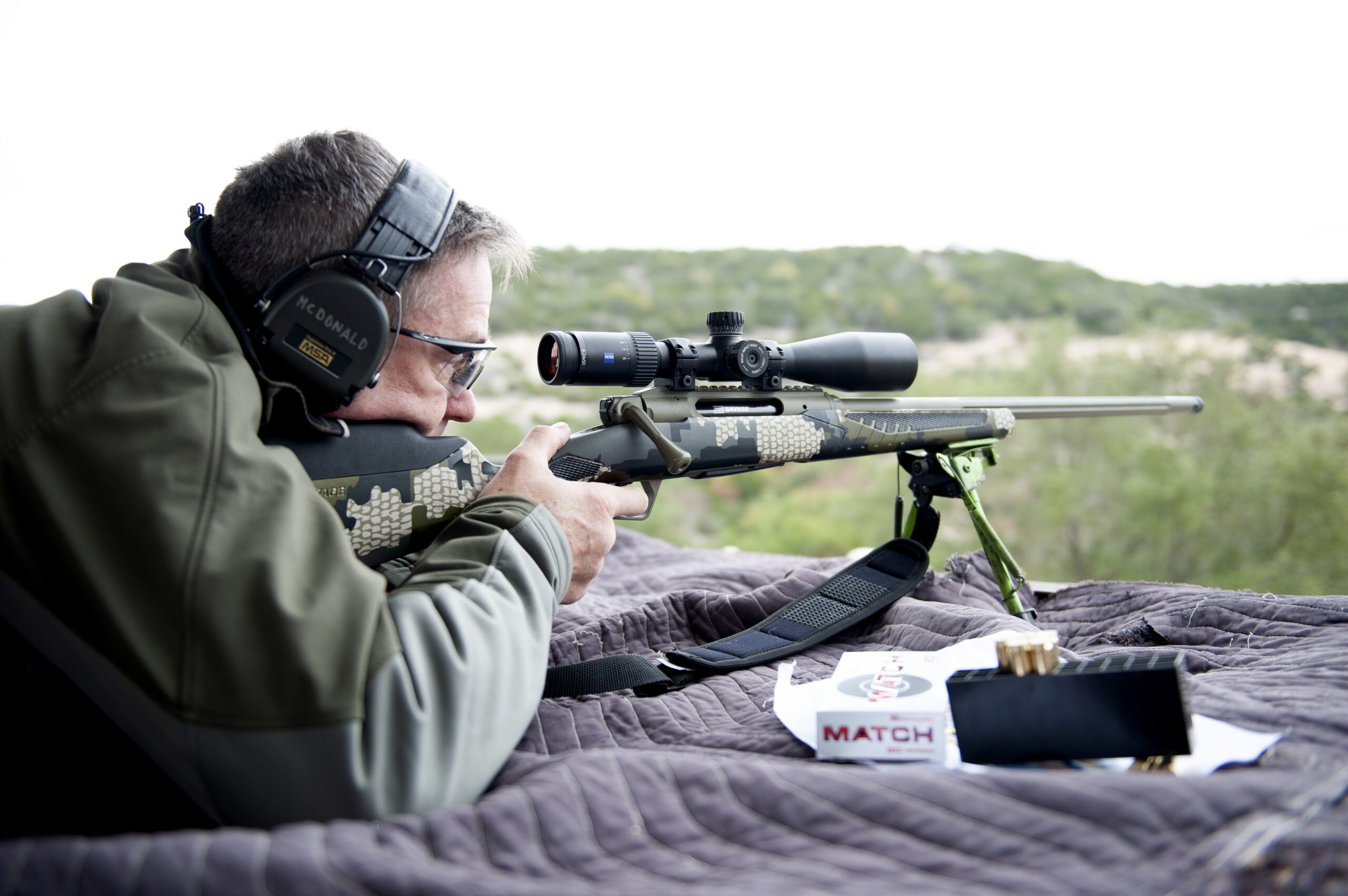 A man with glasses and ear protection lying prone behind a camo rifle on a shooting blanket in rugged country.