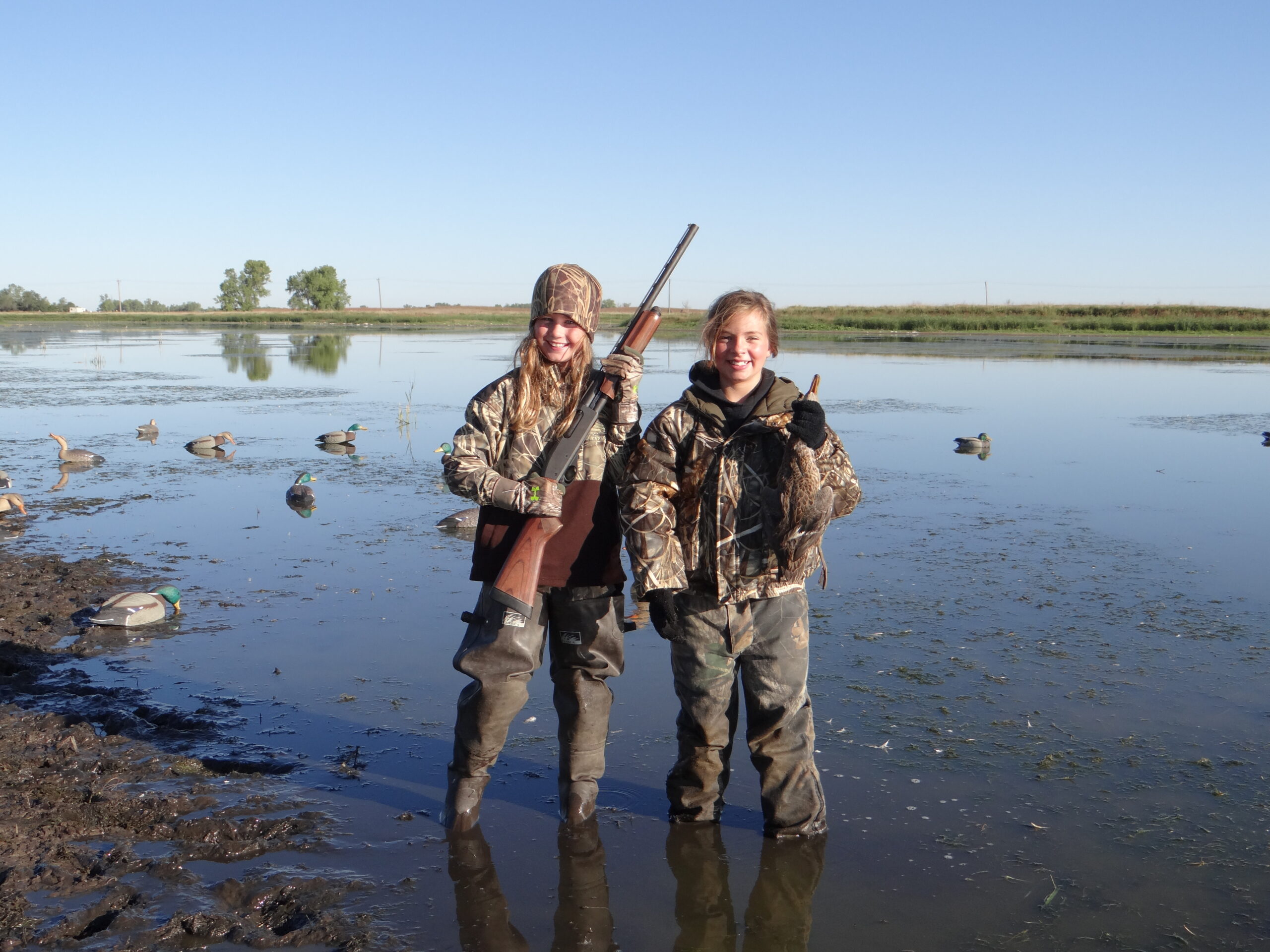 A pair of youth waterfowl hunters hold up a shotgun and a duck.