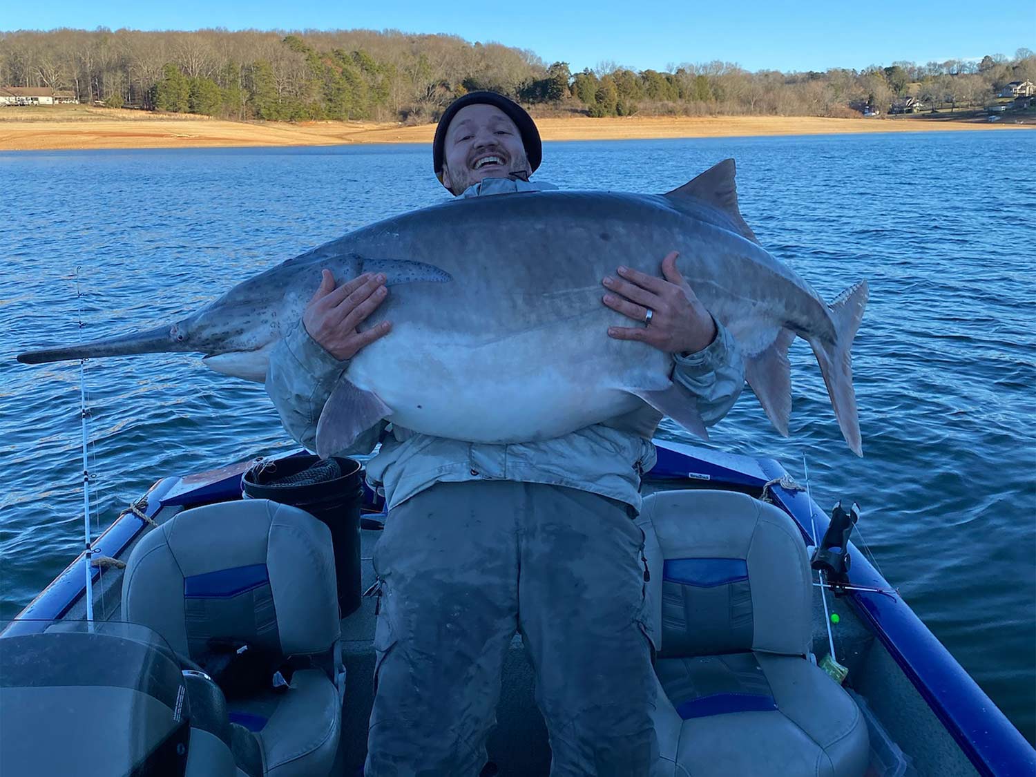 An angler stands in a boat holding up a large paddlefish.