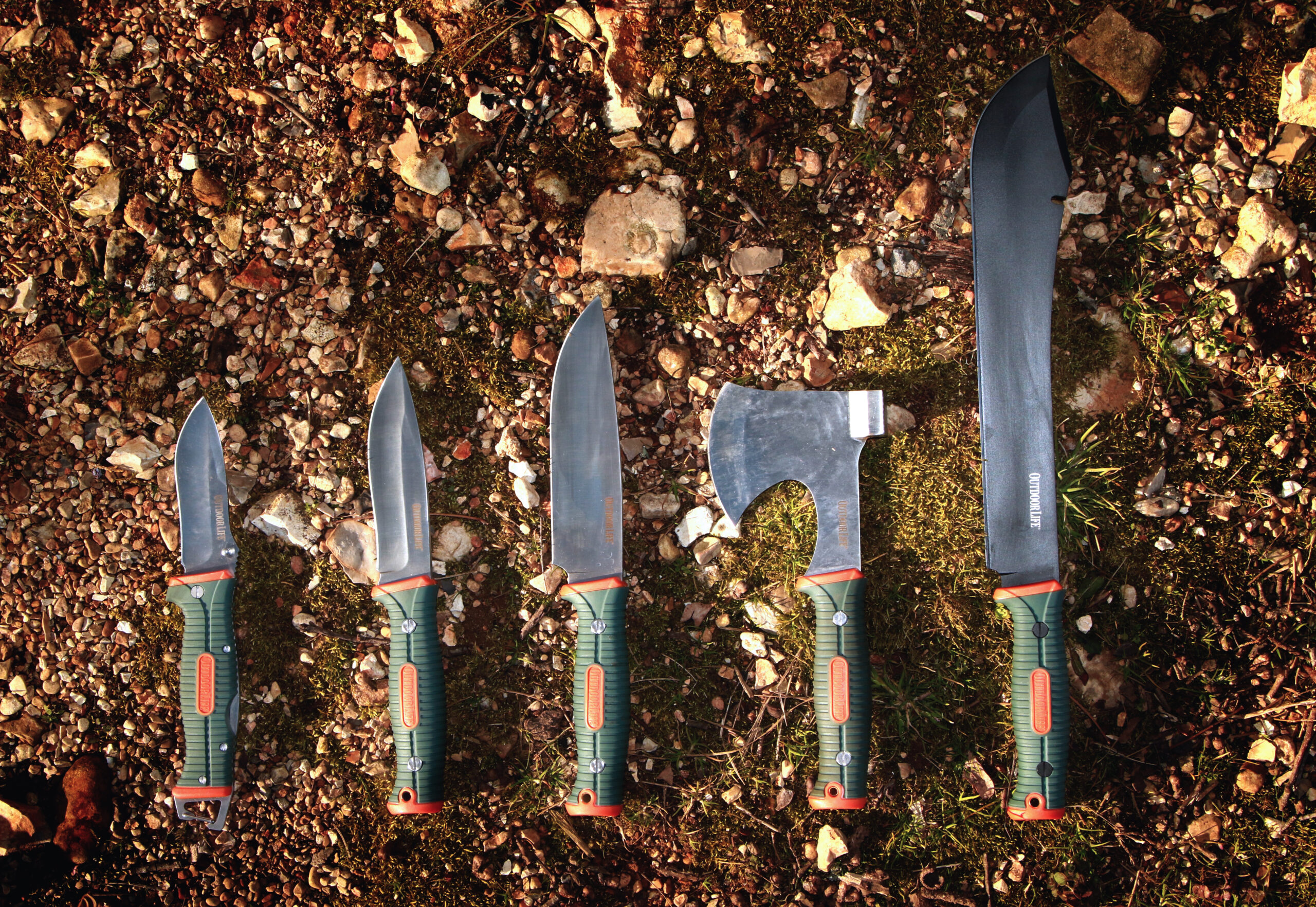 A row of Outdoor Life brand camping tools and knives, on a gravel and moss background.