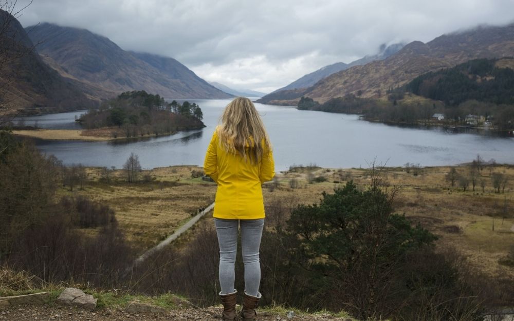 A blonde woman with yellow jacket standing staring the big lake between mountains.