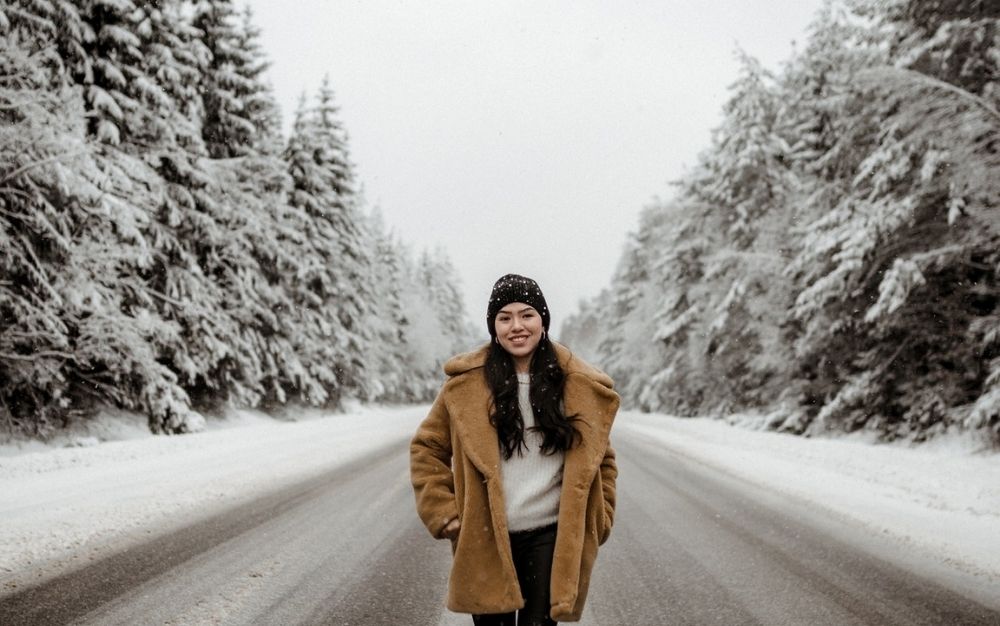 A girl smiling and walking into a snowy road between some trees, while wearing a long and comfortable warm brown jacket.