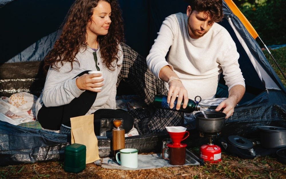 A boy and a girl standing outside and drinking coffee from the thermos while other cups are launched into the ground with a blanket.