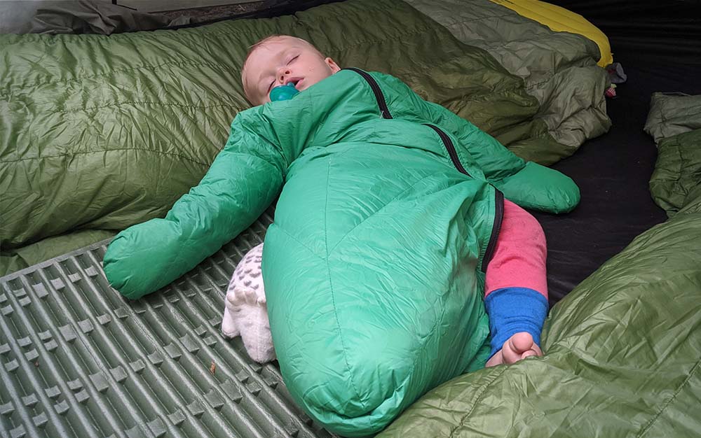 A toddler in a green sleeping bag with arms