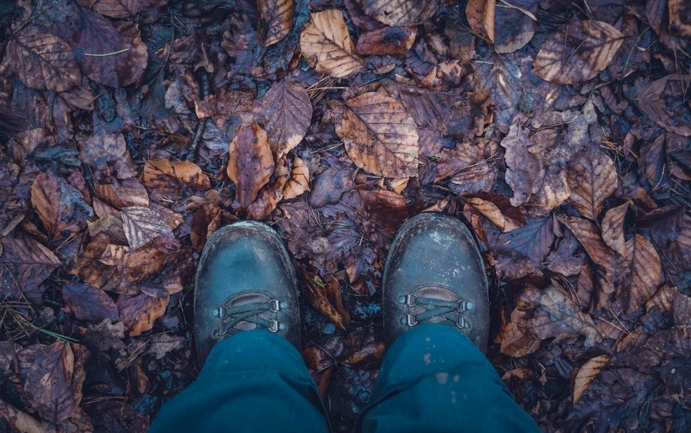 A pair of black boots of a person who treads on a ground with wet soil and leaves.