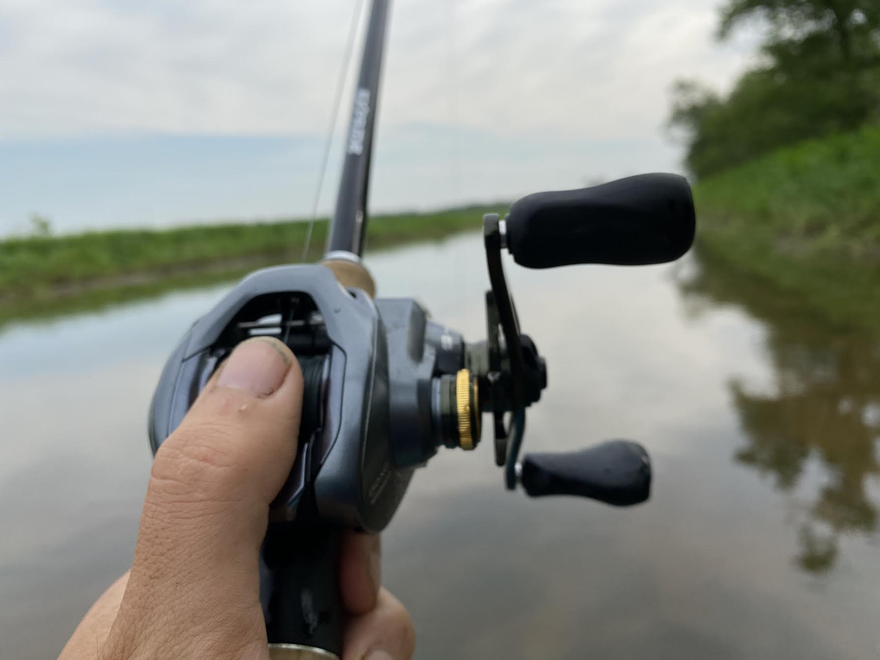 An angler's thumb on a baitcasting reel over a body of water