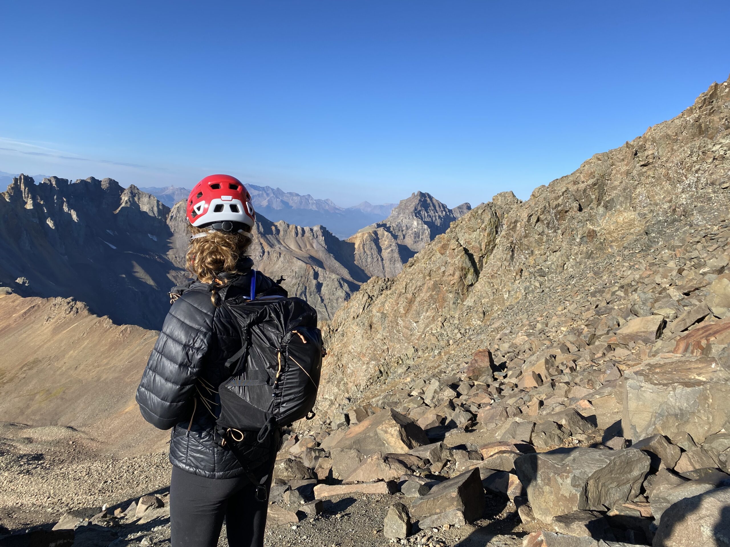 A woman with a black jacket, black backpack, and red climbing helmet looking over mountains