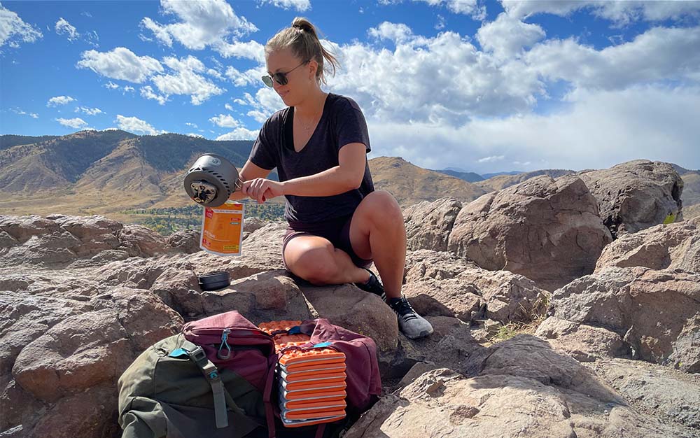 A woman pouring water into an orange bag of dehydrated food sitting on a rock overlooking the mountains