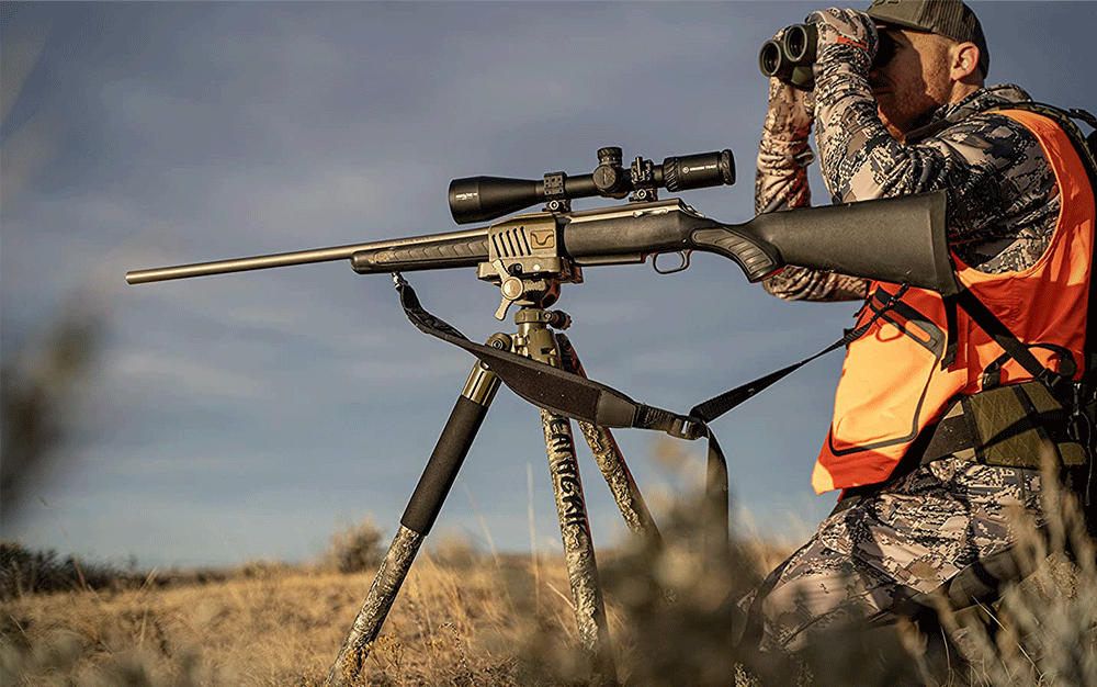 A man in camo and an orange vest using his binoculars next to a rifle on a tripod