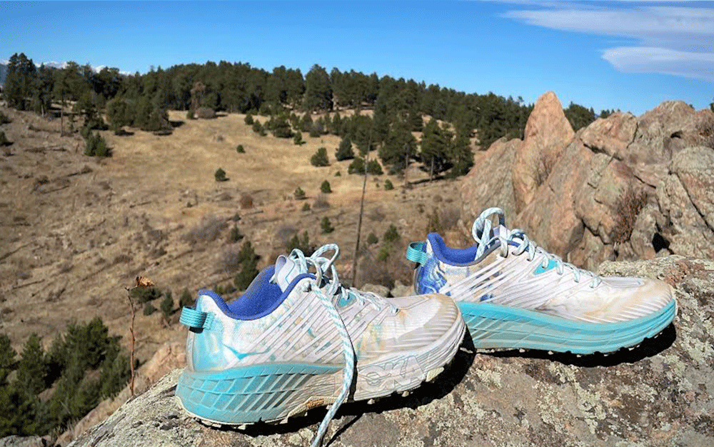 A pair of white and blue Hoka Speedgoat 4s overlooking rocky hills and trees