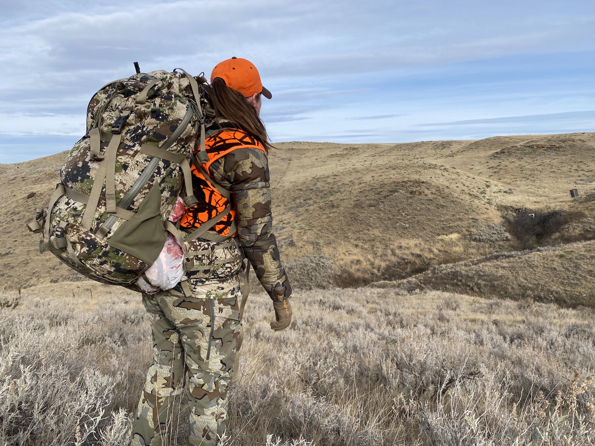 A woman in camo and orange carrying a large camo backpack looking over a field