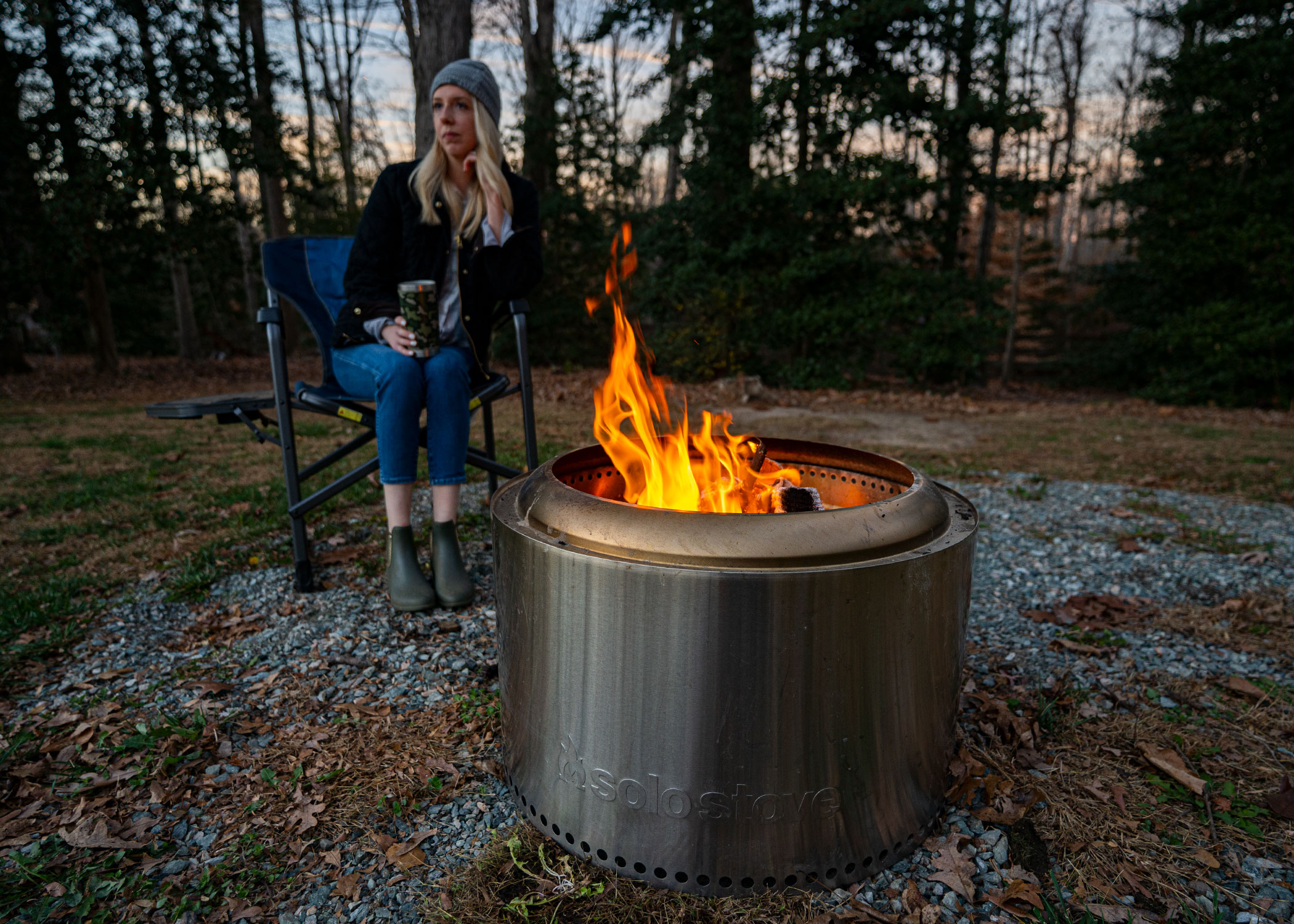 A woman sitting behind a fire pit with trees in the background