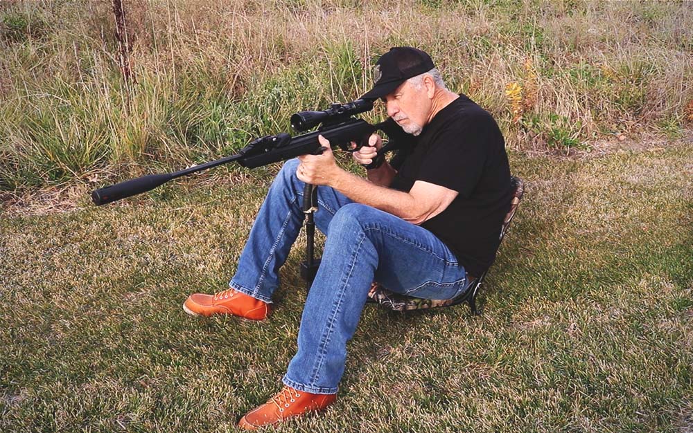 A man sitting on the grass aiming a black air rifle