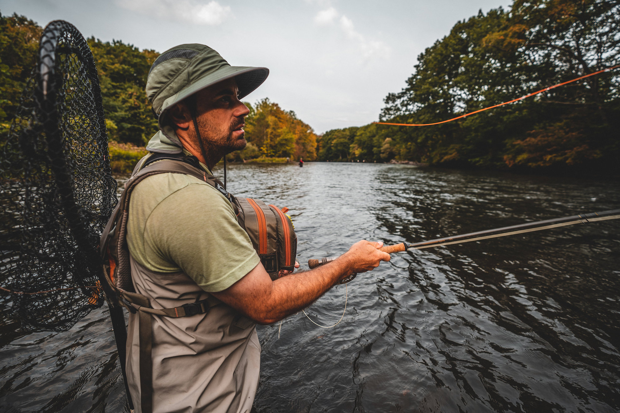 A man fishing on a river wearing a fishing vest and a net on his back