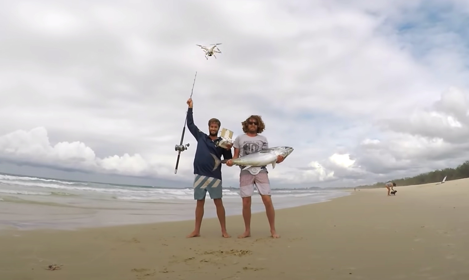 Two fishermen on the beach, one holding a rod, the other a fish, with a drone above them.