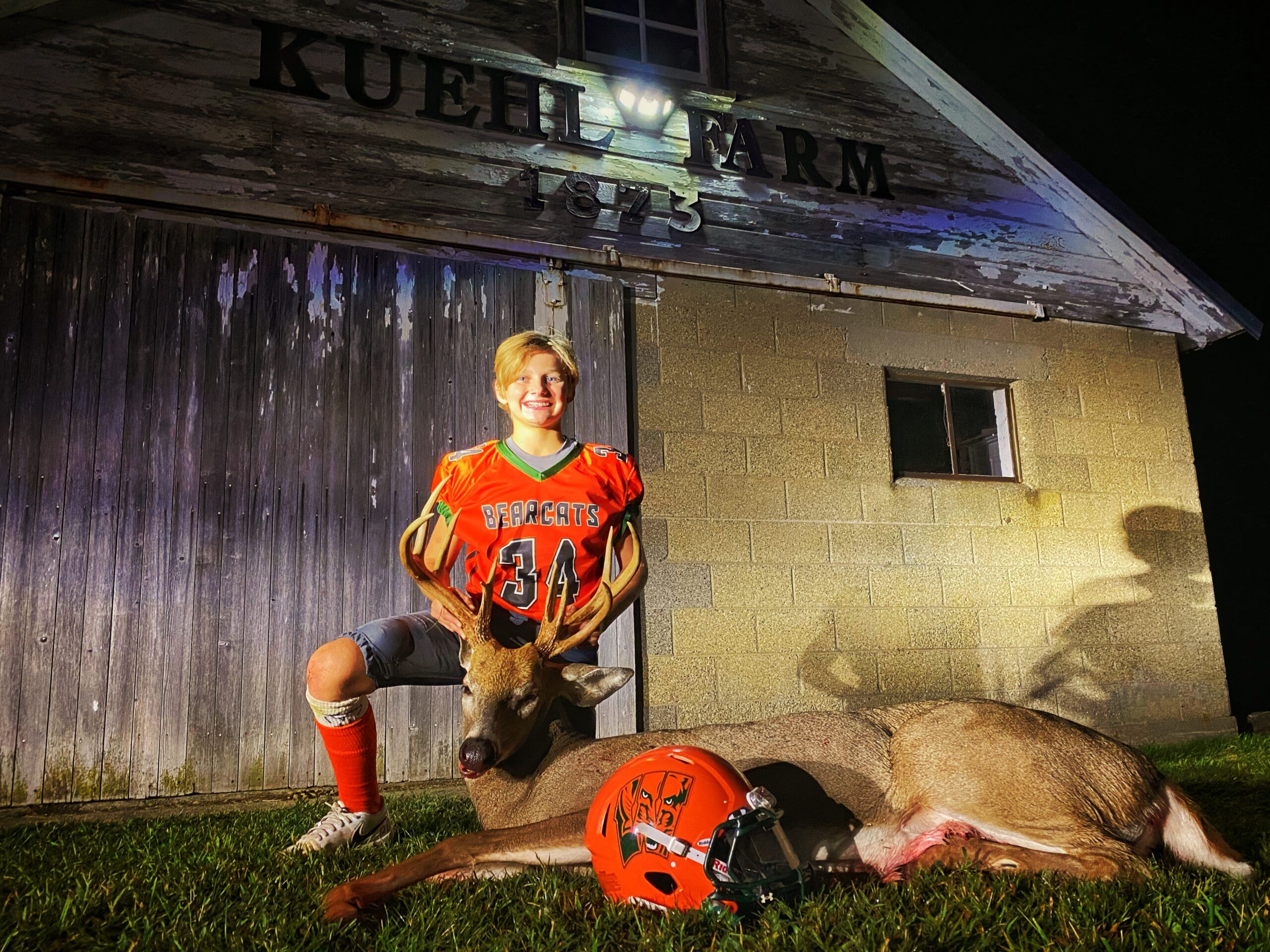 A young deer hunter in a football uniform kneels beside his buck.