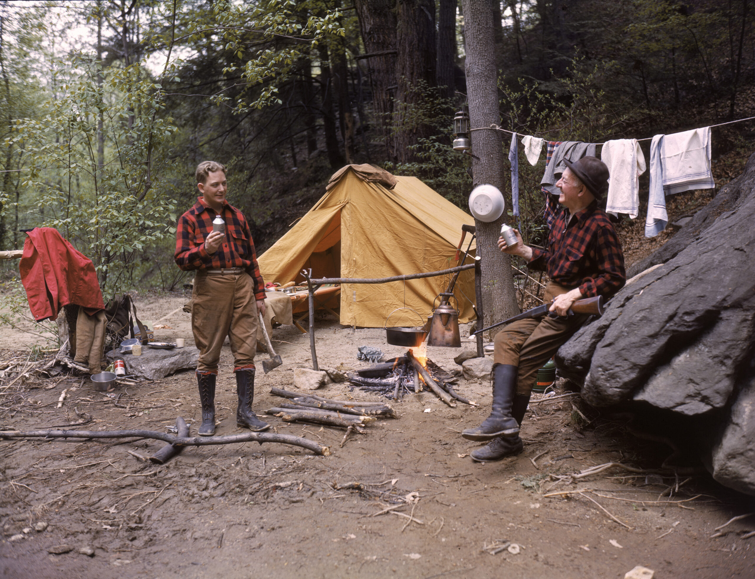 Two hunters in red plaid shirts drink beer in front of a wall tent camp.