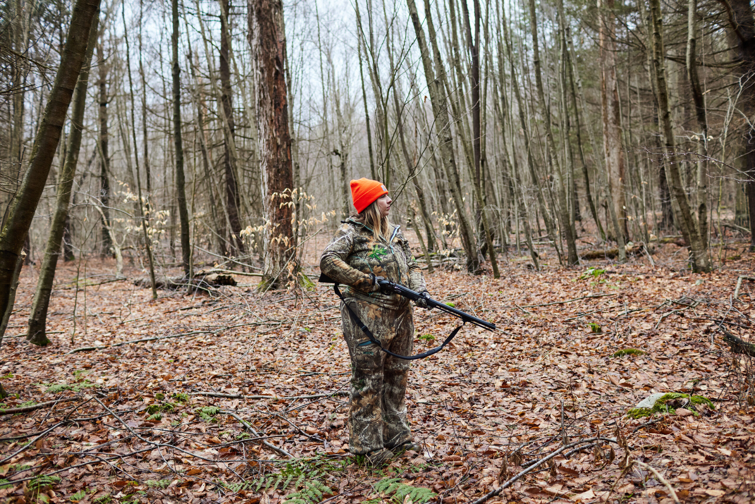 A hunter in her mid-20s stands with a flintlock rifle, gazing into the woods.