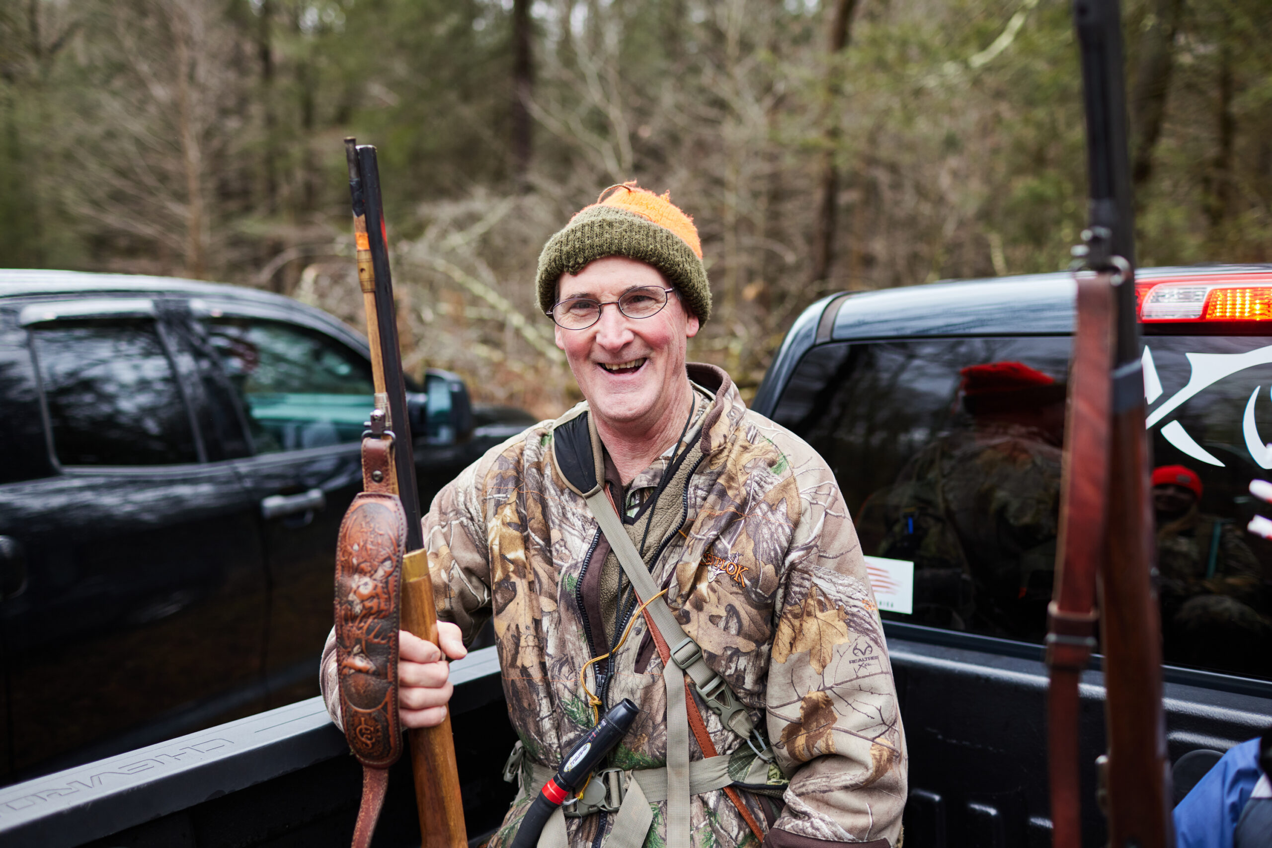 A smiling hunter stands by a pair of pickups, holding a flintlock rifle.