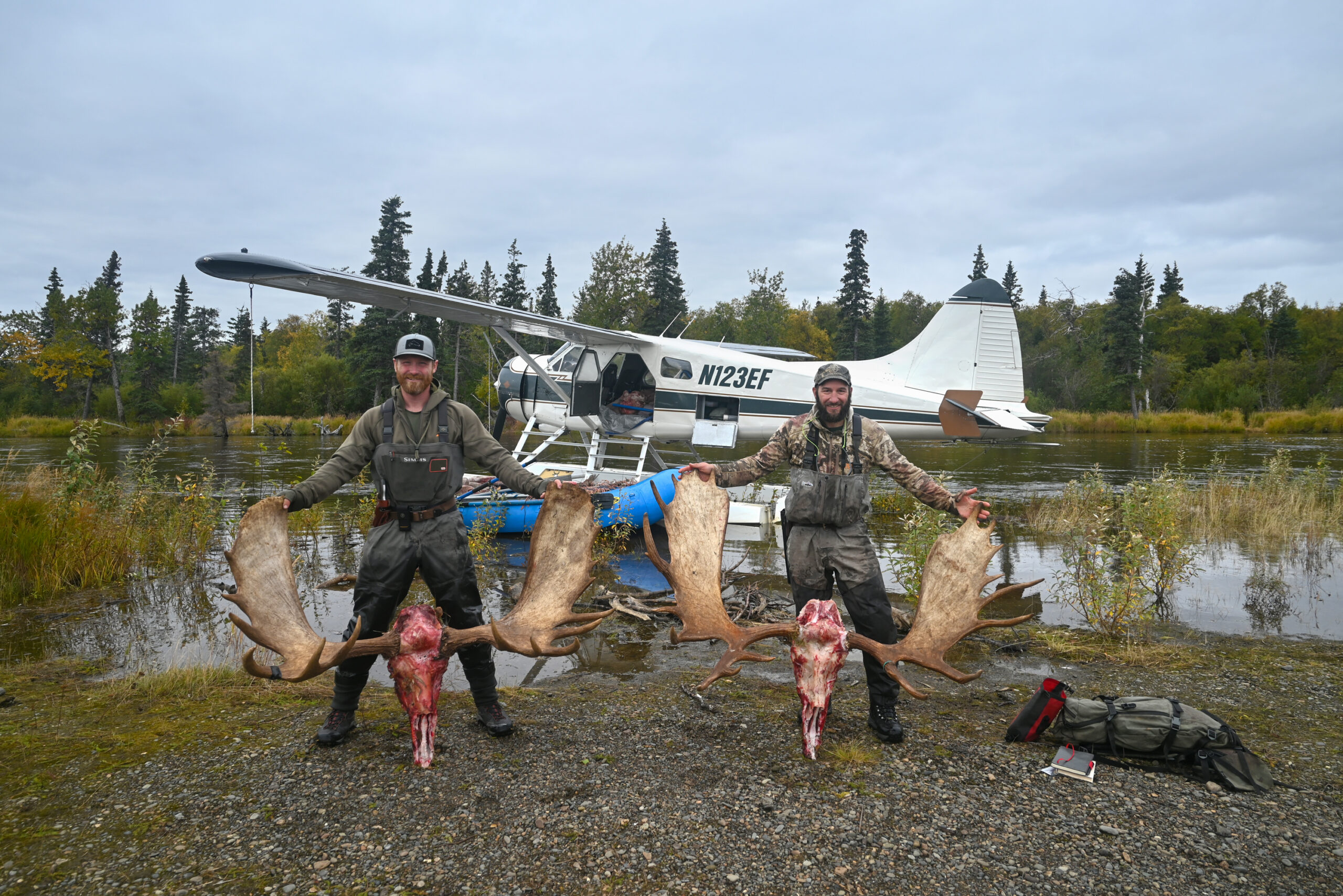 Two hunters stand with their moose skulls in front of a float plane.